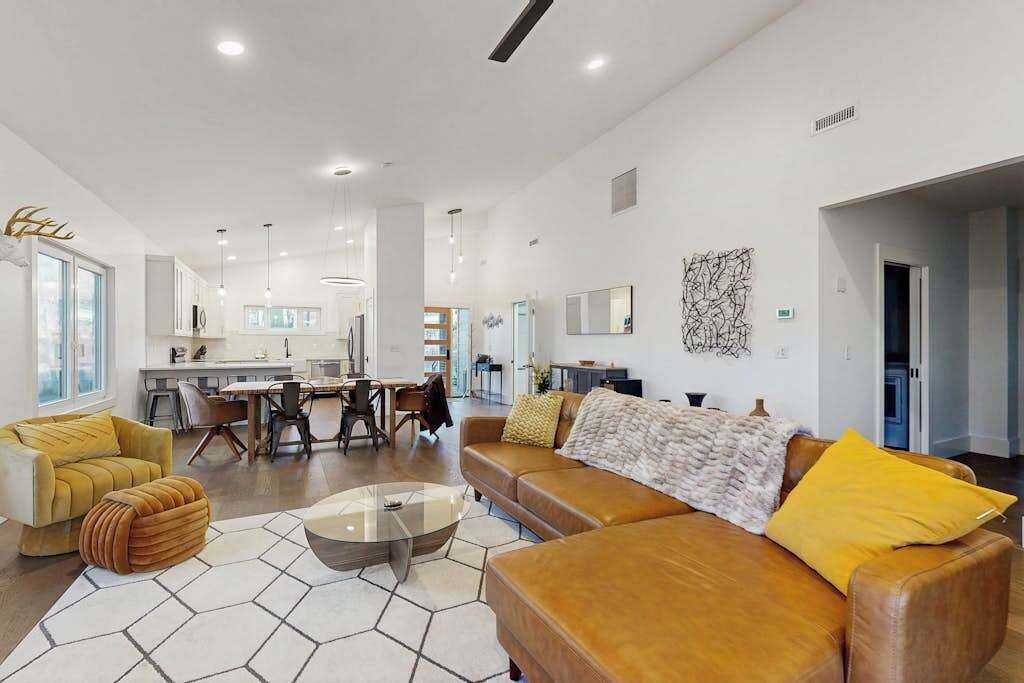 A spacious living area with a sofa, ottoman, and coffee table, a dining area with chairs, and a kitchen in the background.
