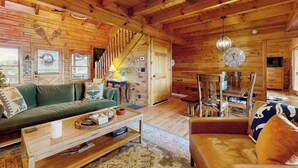 Interior de una cabaña de madera con sofá, mesa de centro, mesa de comedor, sillas y una escalera.