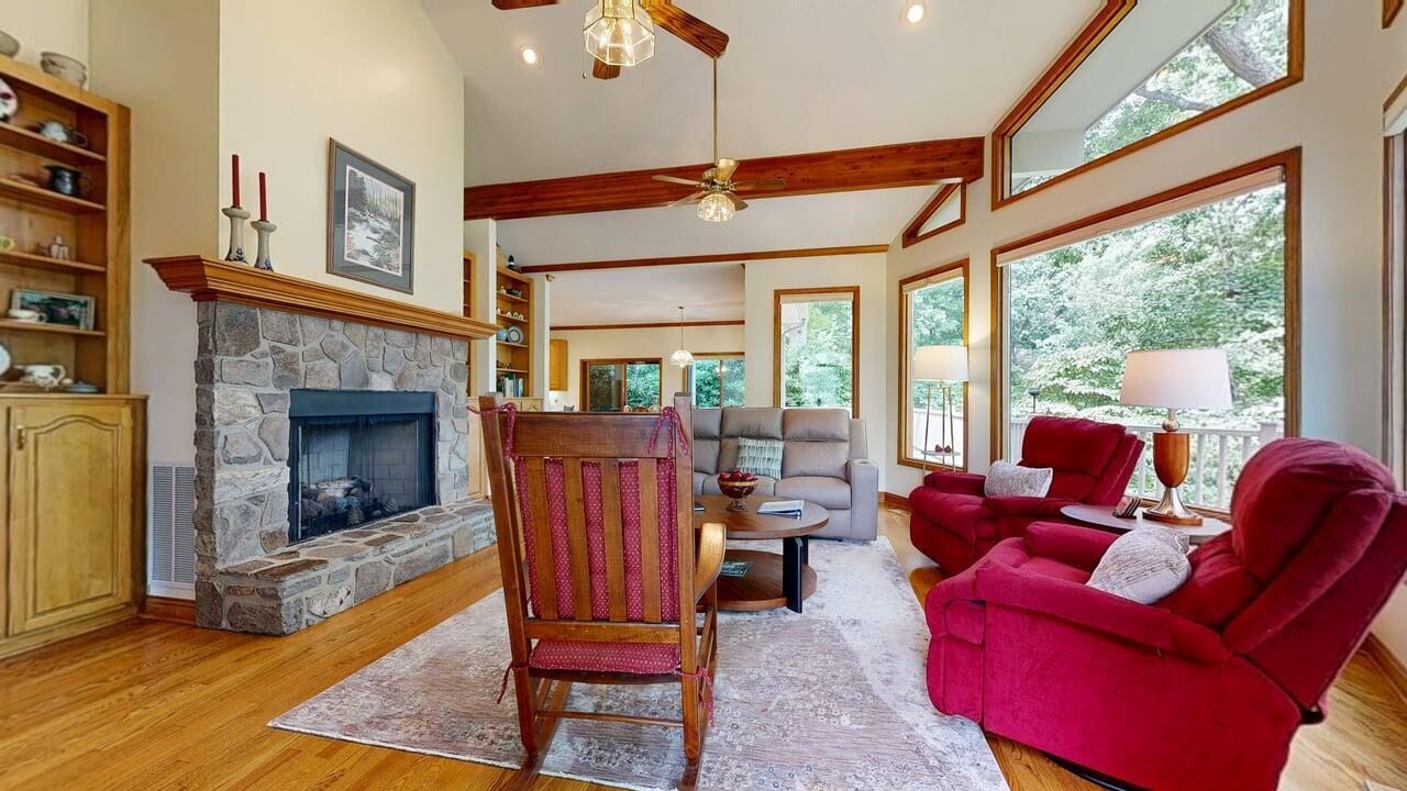 A cozy living room with a stone fireplace, wooden beams, a red armchair, a grey sofa, and large windows.