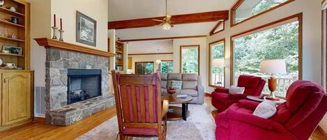 A cozy living room with a stone fireplace, wooden beams, a red armchair, a grey sofa, and large windows.