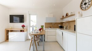 A modern kitchen with white cabinetry, a dining area with a table and chairs, a large clock on the wall, and a TV mounted on the wall.