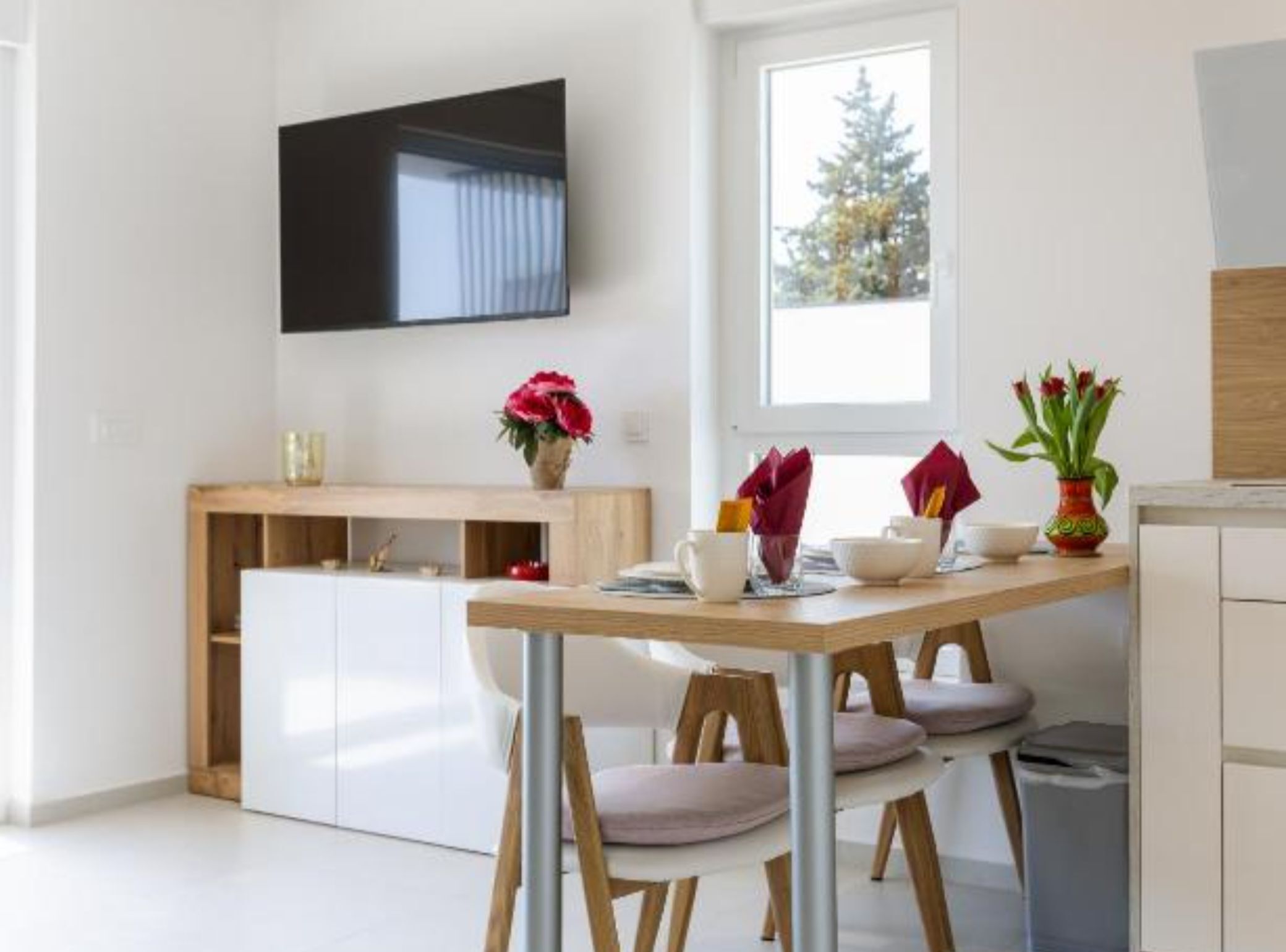 Eine moderne Küche mit Essbereich, einem an der Wand befestigten Fernseher und einem Fenster mit Blick auf Bäume.