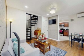 A modern living room with a sofa, a coffee table with flowers, a dining area with chairs, a kitchen with cabinets, and a staircase.