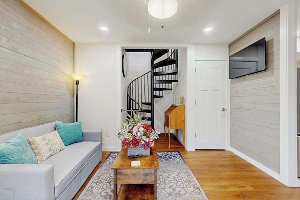 A modern living room with a sofa, a coffee table with a floral arrangement, a staircase, and a TV mounted on the wall.