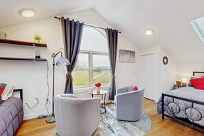 A bedroom with a bed, two chairs, a small table, a floor lamp, and a window with curtains.