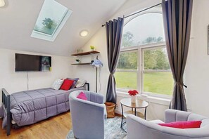 A compact room with a bed, two chairs, a small table, a TV, a window with curtains, and a skylight.