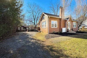 A single-story brick house with a front porch, a chimney, and a gravel driveway. - Centrally located home, with cottage, in Brevard - close to outdoor activities! (Brevard)