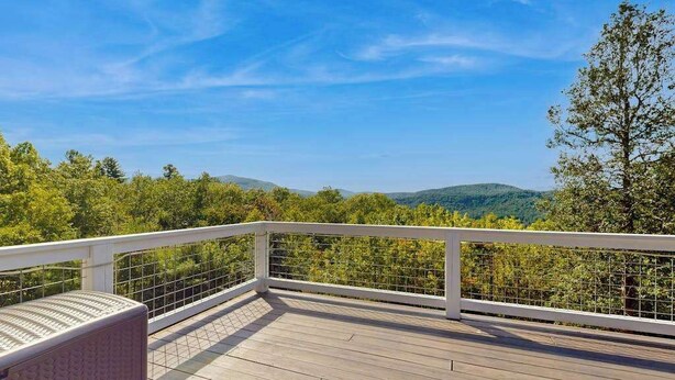 Une terrasse en bois avec une balustrade blanche, donnant sur une zone boisée avec des collines à l’horizon.