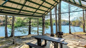 A covered picnic area with a table and benches, overlooking a lake with trees. - Charming house in Brevard with heated outdoor pool, tennis court, and a gym (Brevard)