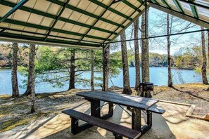 A covered picnic area with a table and benches, overlooking a lake with trees. - Charming house in Brevard with heated outdoor pool, tennis court, and a gym (Brevard)