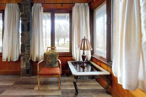 A room with a stone pillar, a chair with a patterned cushion, a table with a lamp, and large windows with curtains. - Morris Villa Lower (Lake Junaluska)