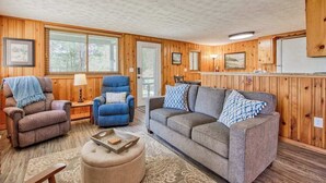 A cozy room with wooden walls, a grey sofa, two armchairs, a small ottoman, and a wooden cabinet.