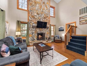 A living room with a stone fireplace, a sofa, a coffee table, and a staircase. - Family Escape at Rumbling Bald with Firepit, Games & Fiber Internet (Lake Lure)