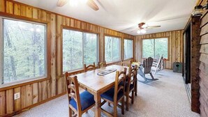 Ein Zimmer mit Holzwänden, einem Esstisch, Stühlen und großen Fenstern mit Blick auf einen Wald.