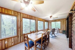 Ein Zimmer mit Holzwänden, einem Esstisch, Stühlen und großen Fenstern mit Blick auf einen Wald.
