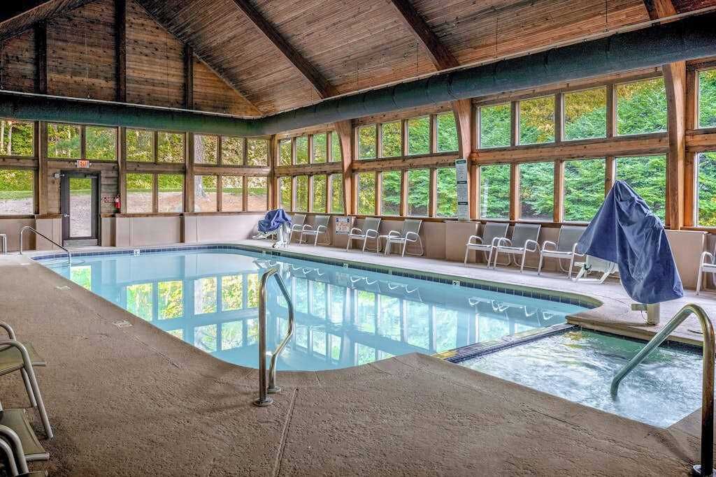 An indoor swimming pool with a view of trees through large windows, a covered seating area, and a wooden ceiling.