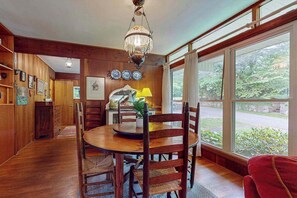 A dining area with a wooden table and chairs, a chandelier, and large windows with a view of greenery. - Friday Cottage (Lake Junaluska)