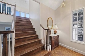 Eine Holztreppe mit weißen Geländern, ein Spiegel, eine Lampe und ein Fenster mit Blick auf Bäume.