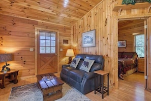 A wooden cabin interior with a leather sofa, a wooden table, a bed, and a window with blinds. - Campground Cabin (Lake Junaluska)