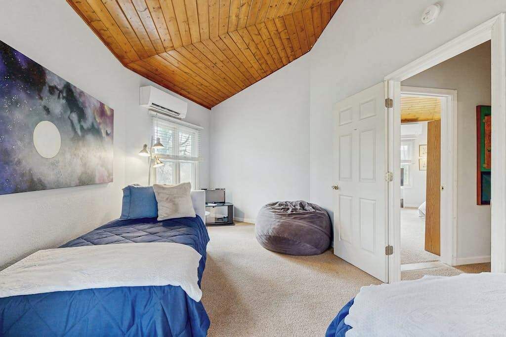 A bedroom with two beds, a wooden ceiling, a wall mural, an air conditioning unit, a bean bag, and a door leading to another room.