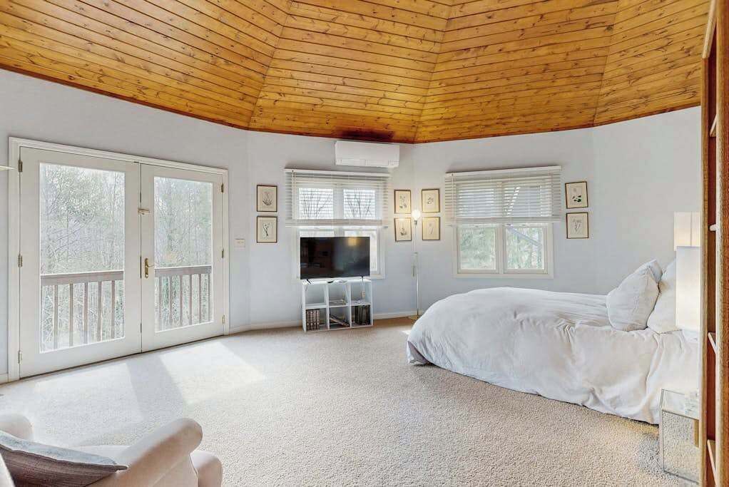 A bedroom with a wooden ceiling, a bed, a TV, and large windows.