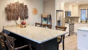 Una cocina con una isla de granito, armarios blancos, un refrigerador de acero inoxidable y un microondas.