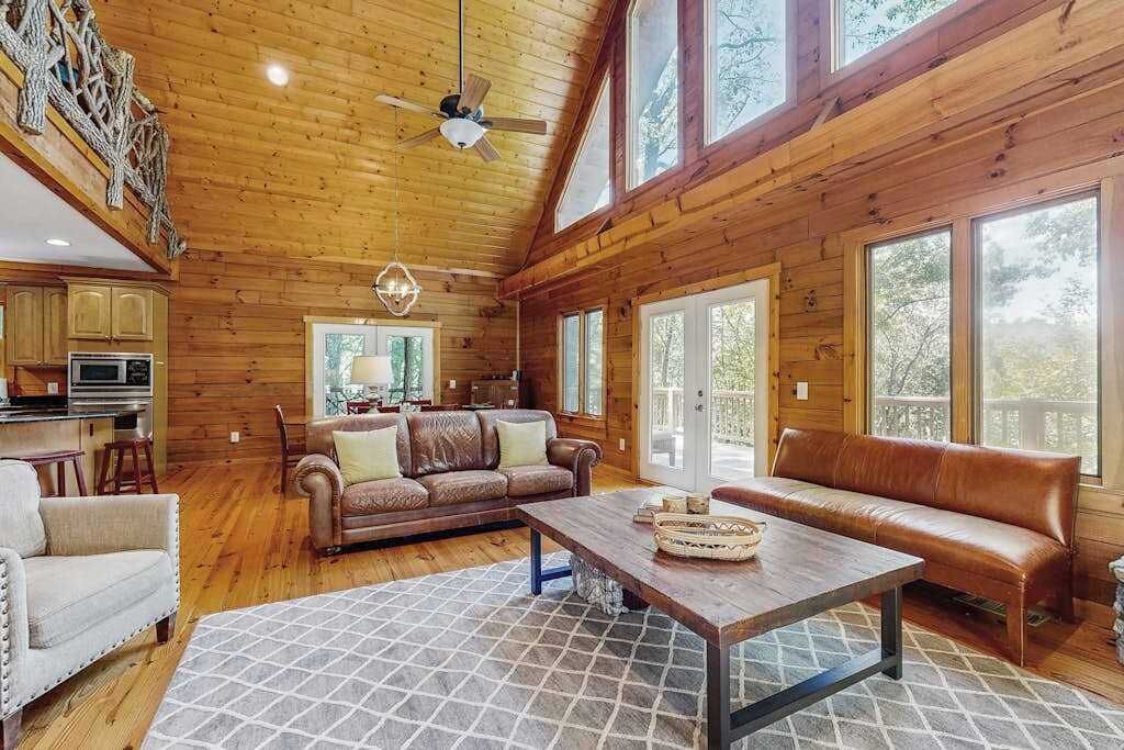 A spacious cabin interior with wooden walls, a large dining table, leather sofas, and a kitchen area.