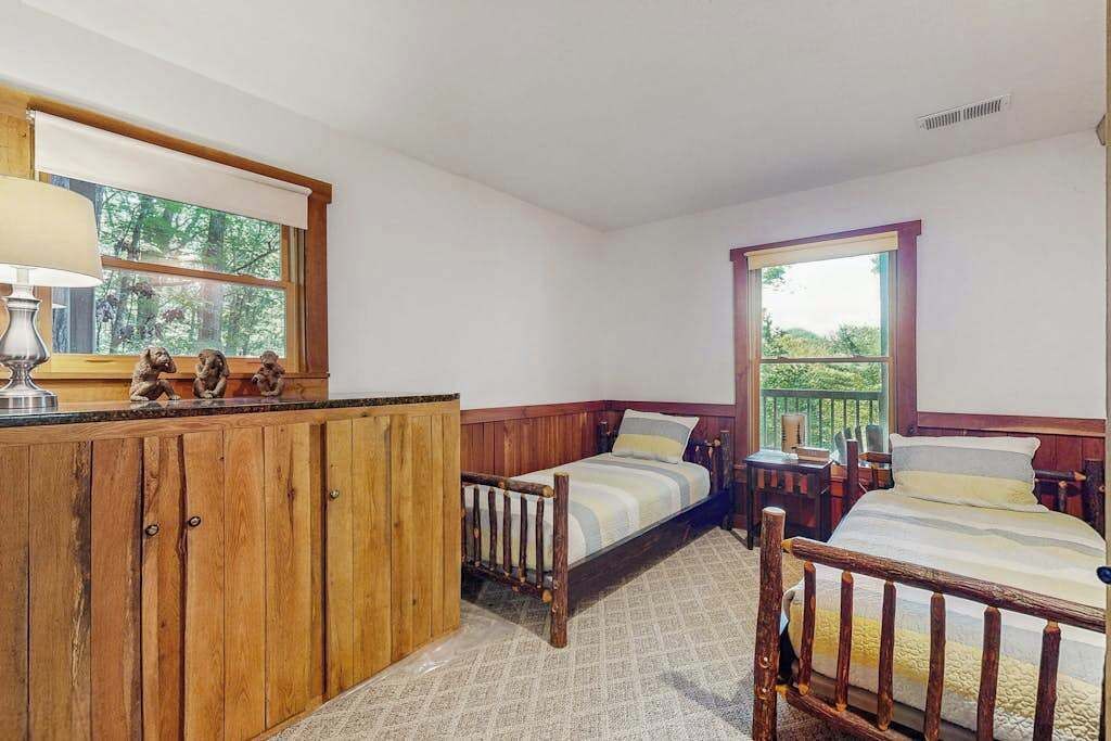Quarto com armário de madeira, duas camas, uma mesa pequena e janela com vista para árvores.