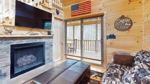 Et sommerhusinteriør med en pejs af sten, et tv ophængt på væggen, en sofa, og et skilt med teksten 'Cabin Sweet Cabin'.