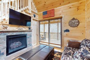 Ein Blockhäuschen-Interieur mit Kamin aus Stein, einem an der Wand befestigten Fernseher, einer Couch und einem Schild mit der Aufschrift 'Cabin Sweet Cabin'.