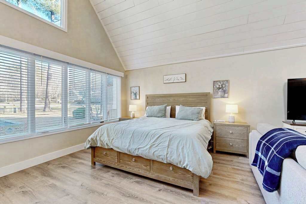 A bedroom with a large bed, a nightstand, a TV, and a window with blinds.