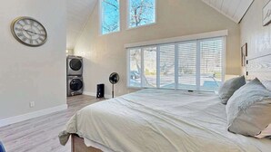 A bedroom with a bed, a clock, a window with blinds, a fan, and a washing machine.