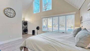 Une chambre à coucher avec un lit, une horloge, une fenêtre avec des stores, un ventilateur et un lave-linge.