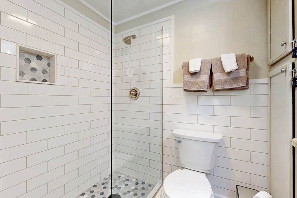 Une salle de bain avec des murs recouverts de carreaux blancs, une douche en verre, des toilettes et deux serviettes accrochées au mur.