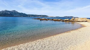 Een strand met helder water, een rotsachtige kustlijn en bergen in de verte.