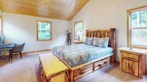 Une chambre avec un plafond en bois, un lit recouvert d’une couverture bleue, un banc en bois, un bureau et une chaise.