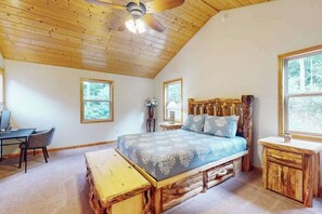 A bedroom with a wooden ceiling, a bed with a blue bedspread, a wooden bench, a desk, and a chair.