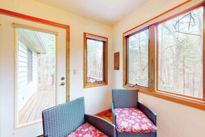 Ein Zimmer mit zwei Korbstühlen, einer Holztür und großen Fenstern mit Blick auf Bäume.
