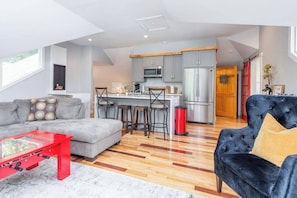 Un espace de vie moderne comprenant un canapé, une table basse rouge, un fauteuil en velours noir, un coin cuisine avec des meubles gris, un réfrigérateur en acier inoxydable et une porte en bois.