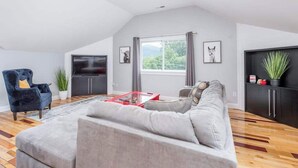 Een moderne woonkamer met een grijze bank, een blauwe fauteuil, een flatscreen televisie en een groot raam met gordijnen.