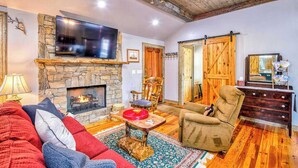 A cozy living room with a stone fireplace, a television mounted above it, a comfortable sofa, and a wooden door. - Stylish dog-friendly cottage with modern feel, fast WiFi, ideal location & more (Swannanoa)