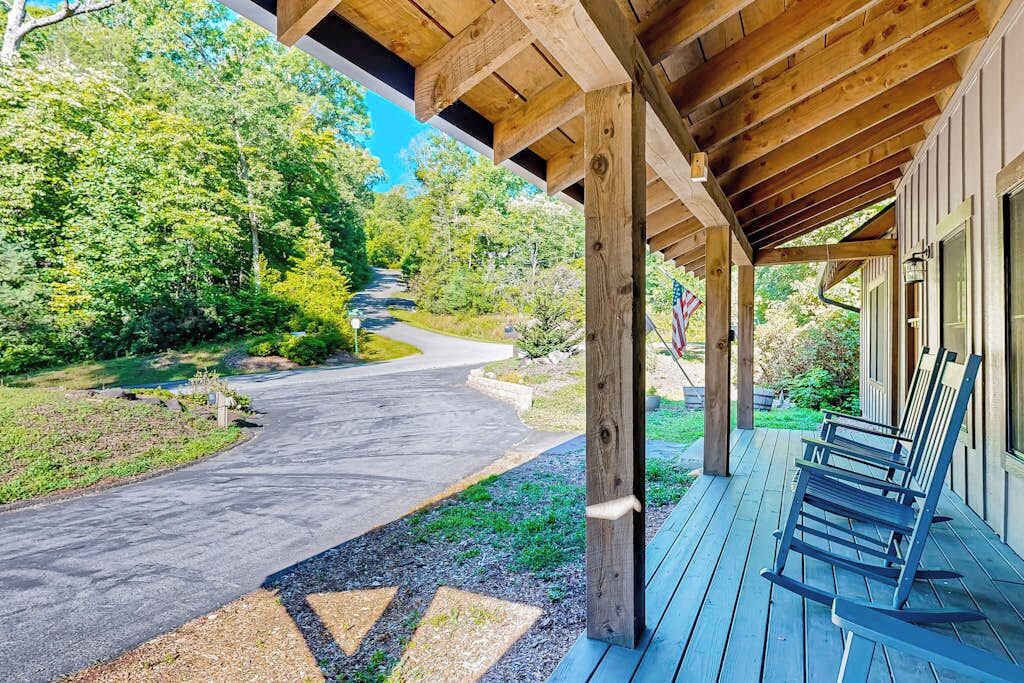 A covered porch with wooden beams, a paved driveway, and a view of a forested area.