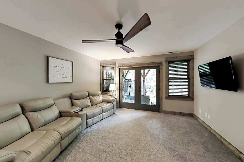 A living room with a beige sectional sofa, a ceiling fan, a framed quote on the wall, and a TV mounted on the wall.