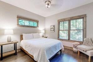 Ein Schlafzimmer mit einem Bett, zwei Nachttischen, einem Sessel, einem Fenster mit Jalousien und einem Deckenventilator.