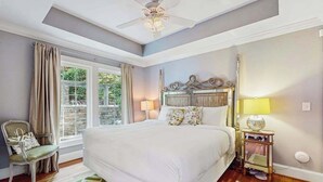 Een slaapkamer met een groot bed, een plafondventilator, een raam met gordijnen, een nachtkastje met een lamp en een stoel.