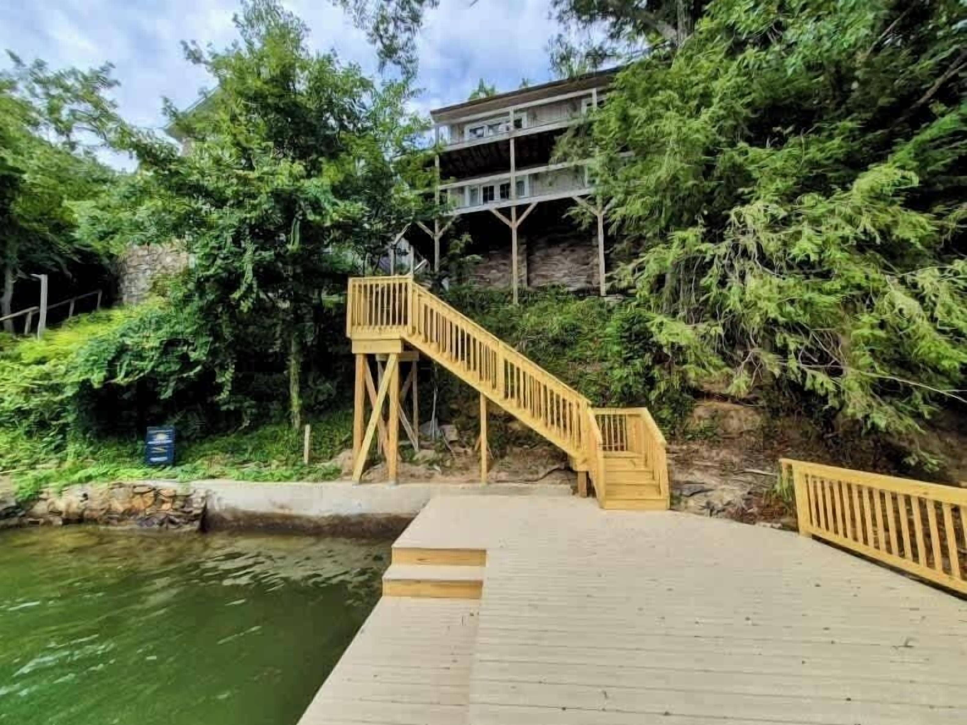 A wooden deck leading to a staircase, surrounded by greenery and a body of water.