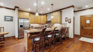 Una cocina con armarios de madera, un refrigerador de acero inoxidable, una isla central con taburetes de bar, iluminación colgante y salpicadero de azulejos.