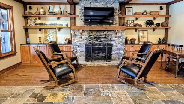 Una chimenea de piedra con un televisor encima, estantes de madera con adornos, y dos mecedoras en una habitación con pisos de madera y vigas a la vista.
