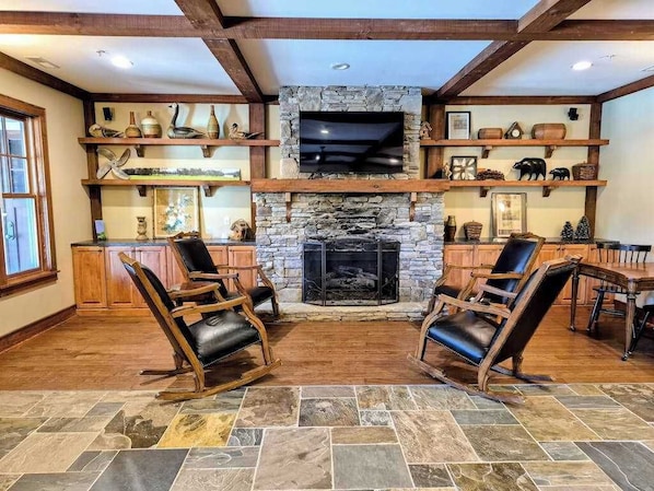 A stone fireplace with a TV above it, wooden shelves with decorative items, and two leather chairs in a room with wooden beams and a stone floor.