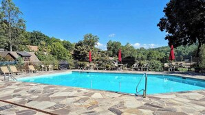 Zona de piscina con baldosas de piedra, sillas de descanso y sombrillas rojas, rodeada de follaje y un cielo despejado.